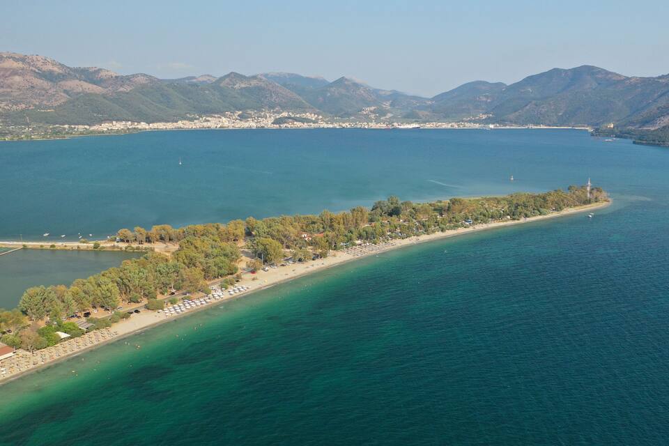An aerial view of a coastal area with a long, narrow beach, a small island with trees and buildings, and mountains in the background.
