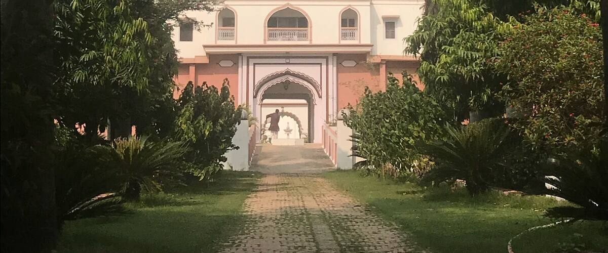 A paved pathway leading to a building with an arched entrance, surrounded by lush greenery and palm trees.