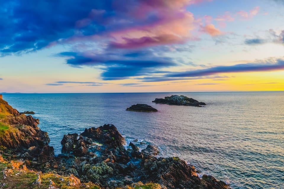 Un paysage côtier avec un phare, des affleurements rocheux et un coucher de soleil sur l’océan.