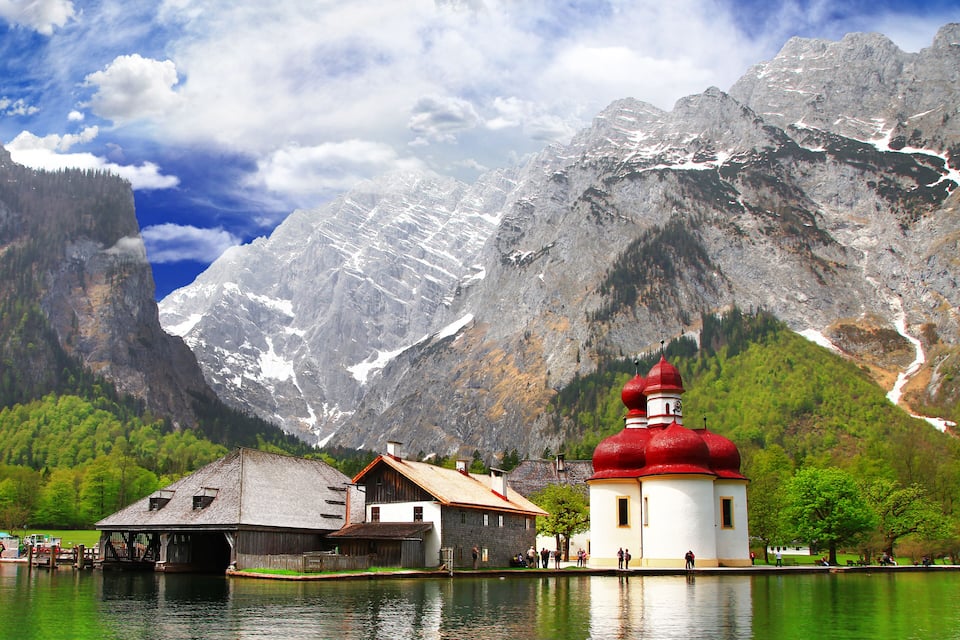 A mountain landscape with a lake, a church with a red dome, and a wooden building by the water.