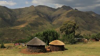 A rural landscape with two thatched-roof huts, a valley, and mountains in the background.