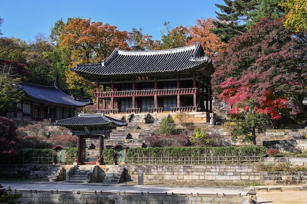Un palais coréen traditionnel, doté d’un toit à plusieurs niveaux, entouré d’un feuillage automnal et de chemins de pierre.