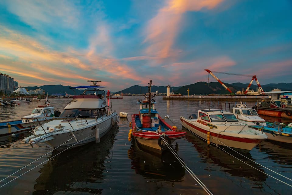 Un port esportiu amb diverses embarcacions amarrades, una grua i muntanyes al fons.