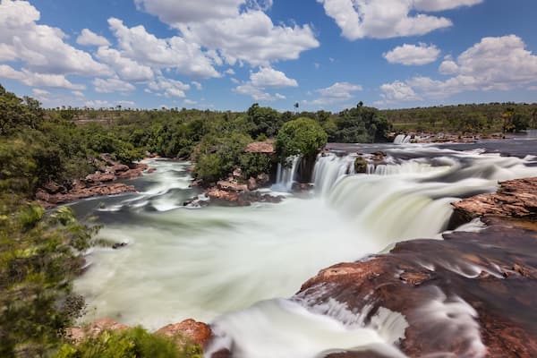 A waterfall cascading over rocks, surrounded by lush greenery and a clear blue sky.