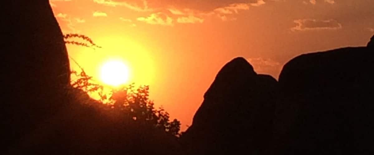 A sunset with the sun partially obscured by clouds, silhouetted mountains, and a tree line.
