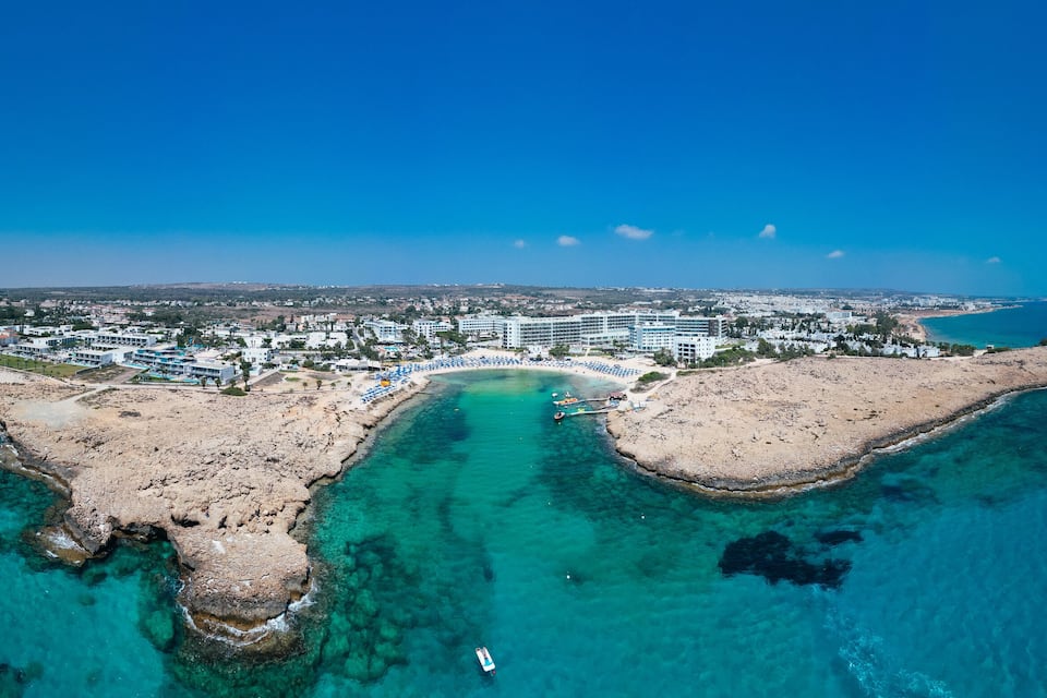 An aerial view of a coastal resort with clear turquoise waters, a sandy beach, and a cluster of hotels.