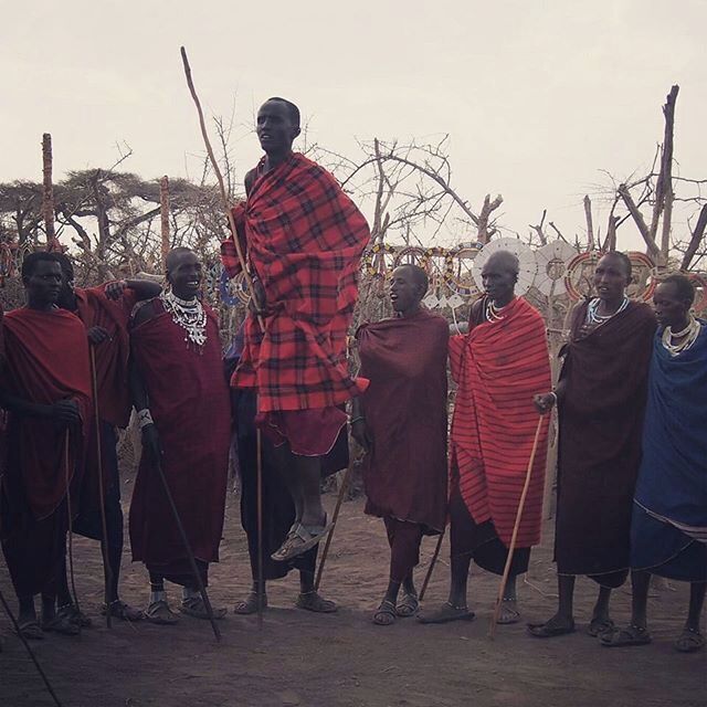 A group of people in traditional attire standing in a natural setting with sparse trees.