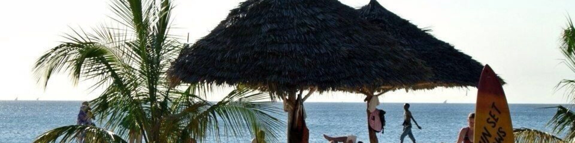 Una playa con una cabaña de techo de paja, palmeras y una tabla de surf con la leyenda 'SUN SET BUNGALOWS'.