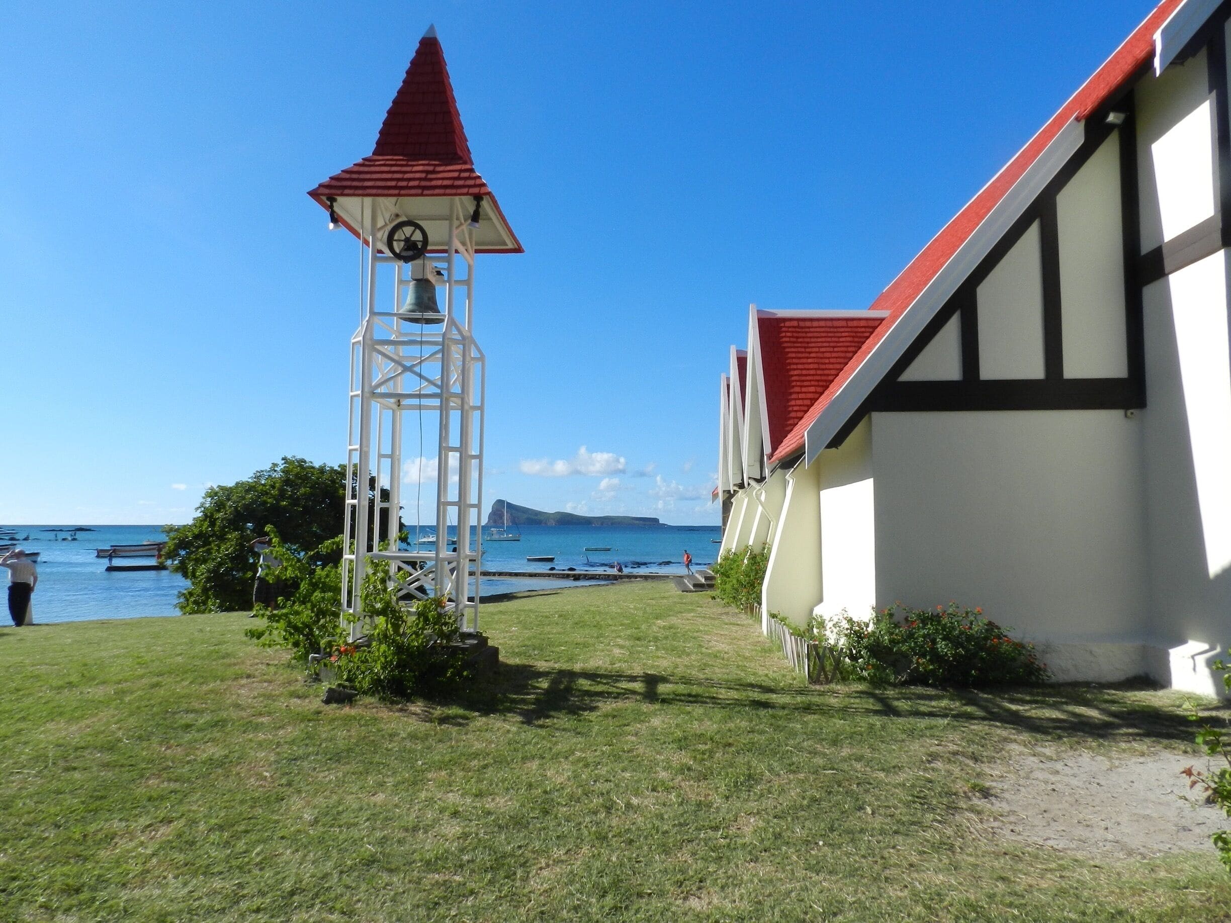 Uma paisagem costeira com uma torre de sino branca, um edifício com telhado vermelho e barcos na água.