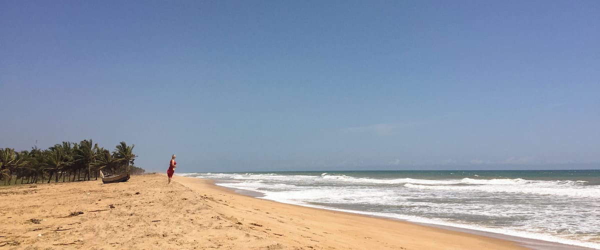 A beach with a person walking, palm trees, and a boat on the sand.