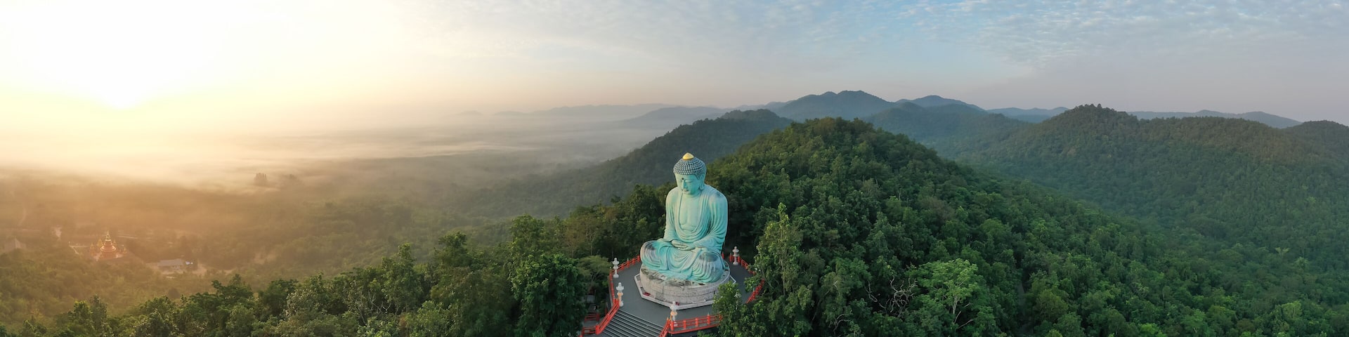 A large Buddha statue on a mountain with a forested valley and misty mountains in the background.