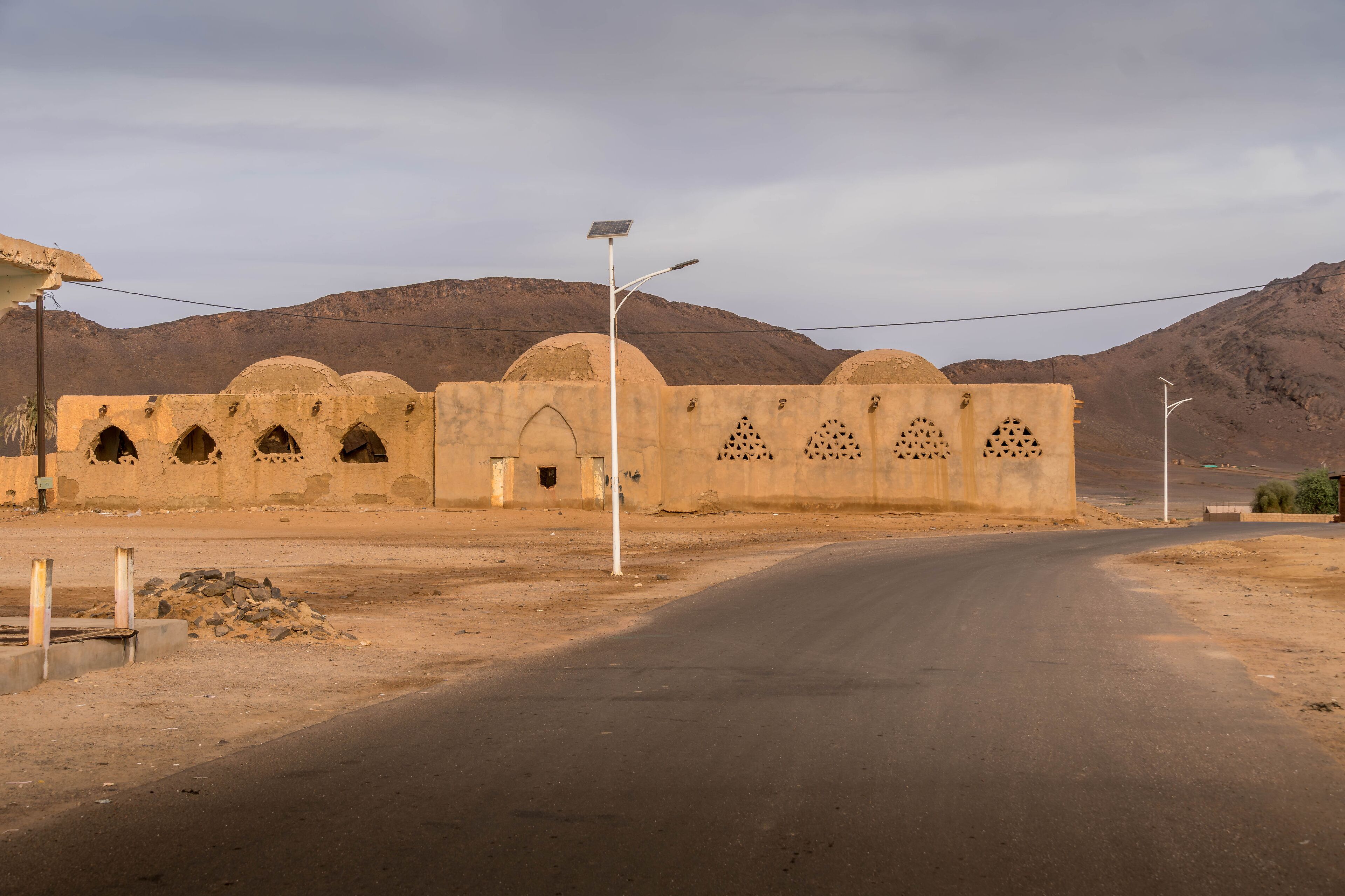 Old mosque with domed roofs and arched windows stands in Fderik, Mauritania, framed by Sahara mountains and empty asphalt road in remote desert town.

