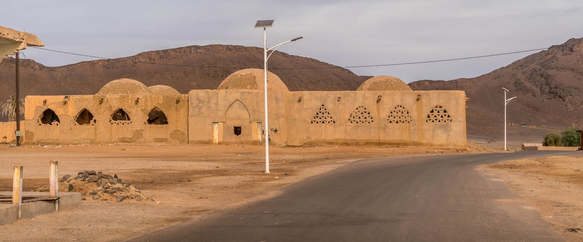Old mosque with domed roofs and arched windows stands in Fderik, Mauritania, framed by Sahara mountains and empty asphalt road in remote desert town.
