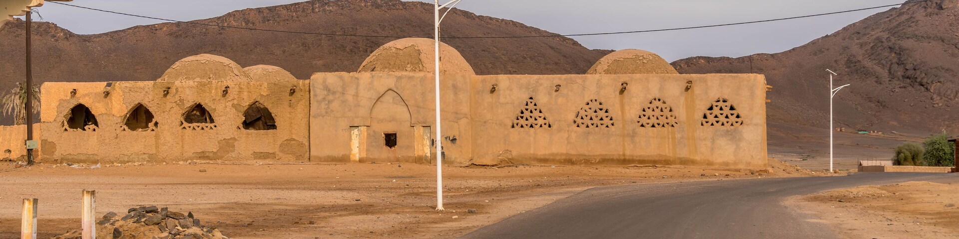 Old mosque with domed roofs and arched windows stands in Fderik, Mauritania, framed by Sahara mountains and empty asphalt road in remote desert town.