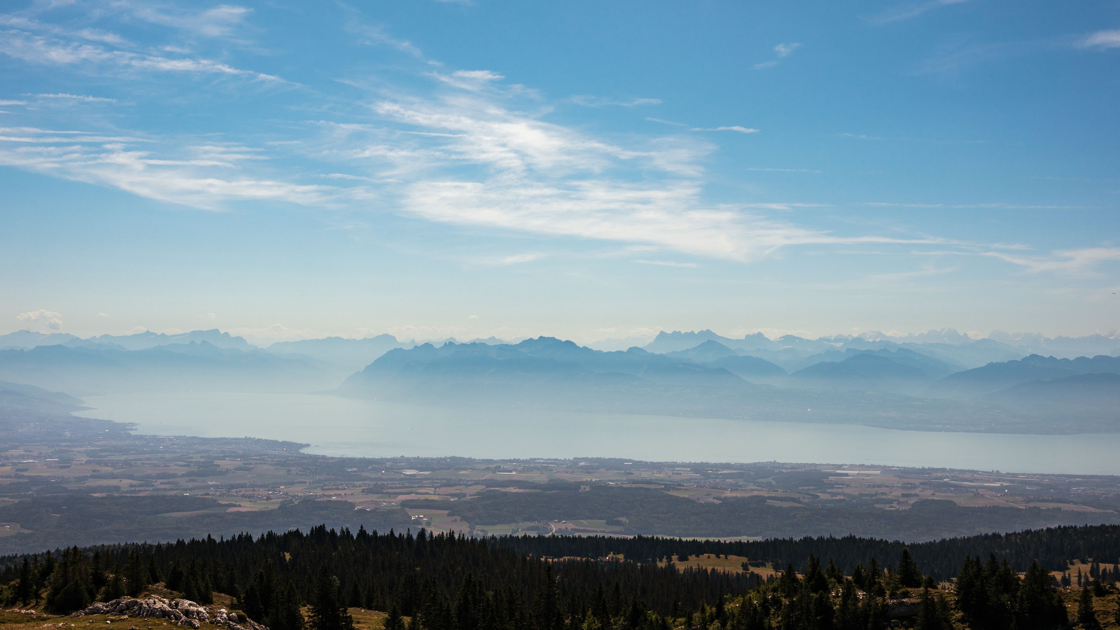 A mountainous landscape with a lake, forested areas, and distant mountain peaks under a clear sky.
