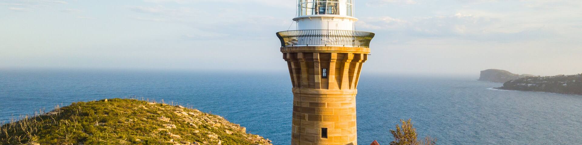 Un faro con techo rojo, cúpula blanca y veleta en la parte superior, con vistas a un paisaje costero bajo un cielo despejado y un mar tranquilo.
