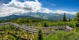 Eine gewundene Holzbrücke in einer bewaldeten Berglandschaft, mit fernen Berggipfeln im Hintergrund.