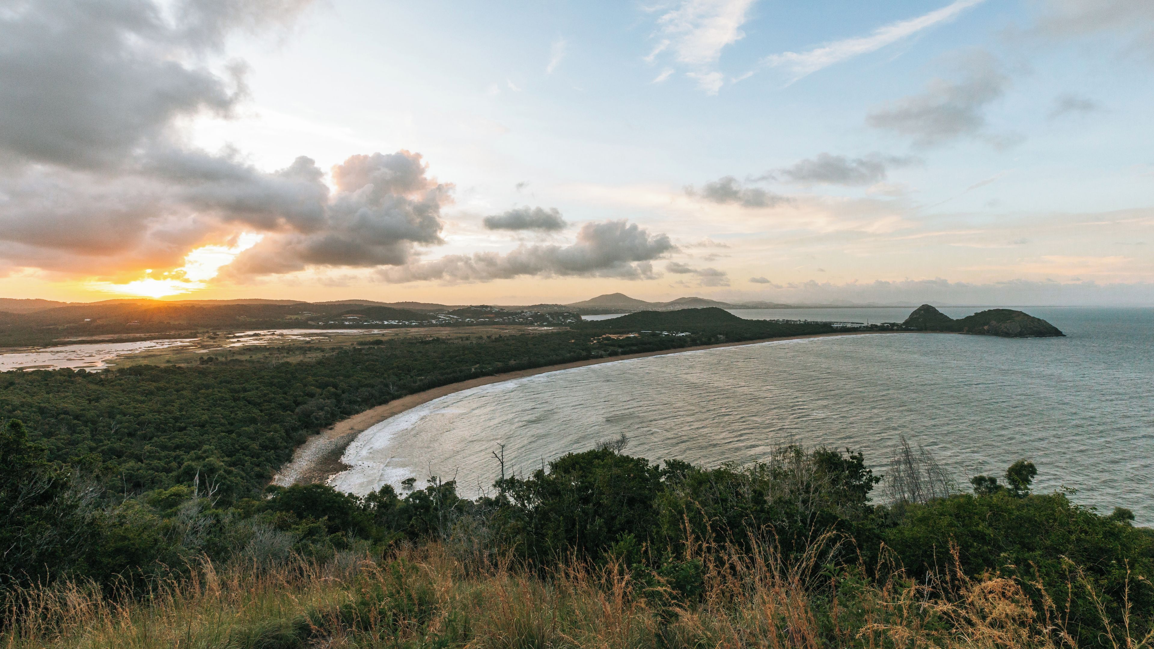 A coastal landscape with a beach, forested hills, and a sunset.