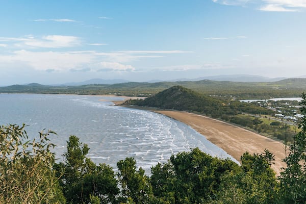 A coastal landscape with a sandy beach, calm sea, and green hills in the background.