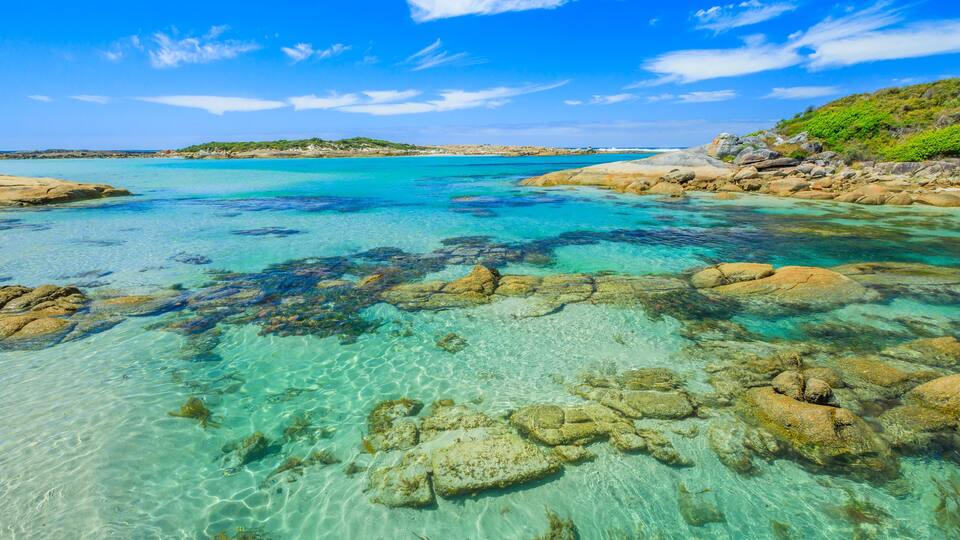 A clear turquoise sea with visible rocks and seaweed, surrounded by a rocky coastline and a distant hill.