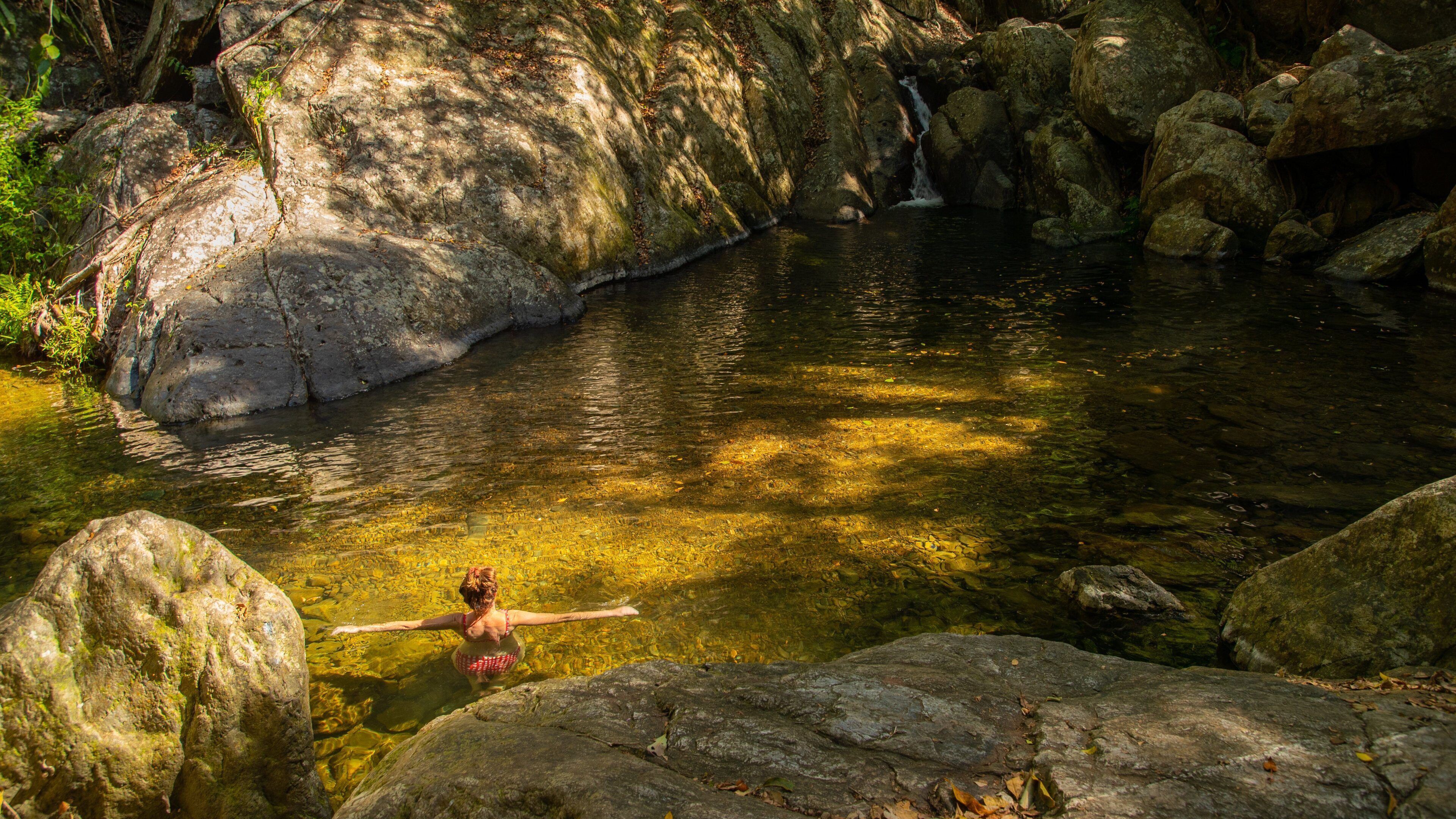 A woman in a red swimsuit standing in a clear stream surrounded by large rocks and trees.