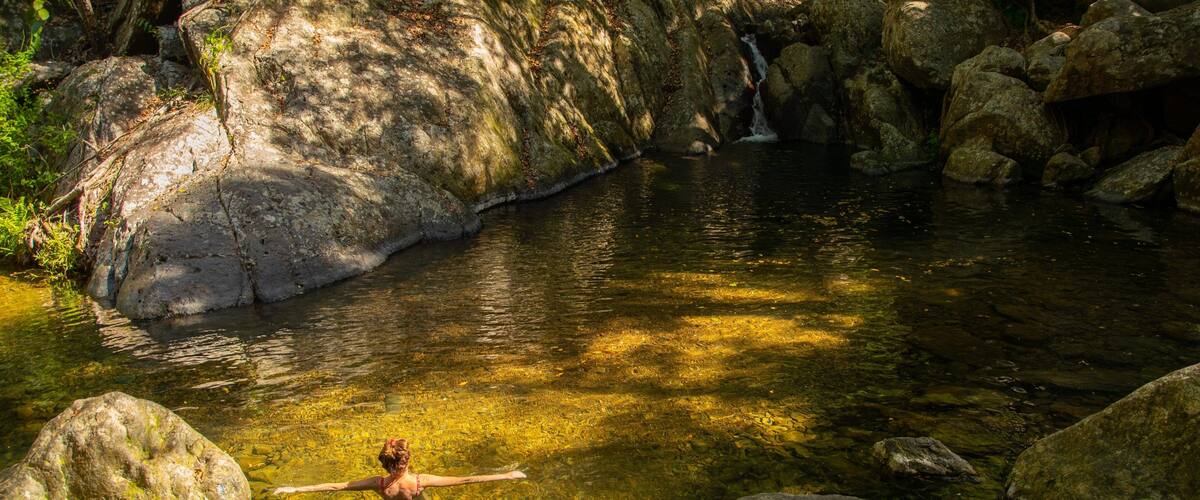 A woman in a red swimsuit standing in a clear stream surrounded by large rocks and trees.