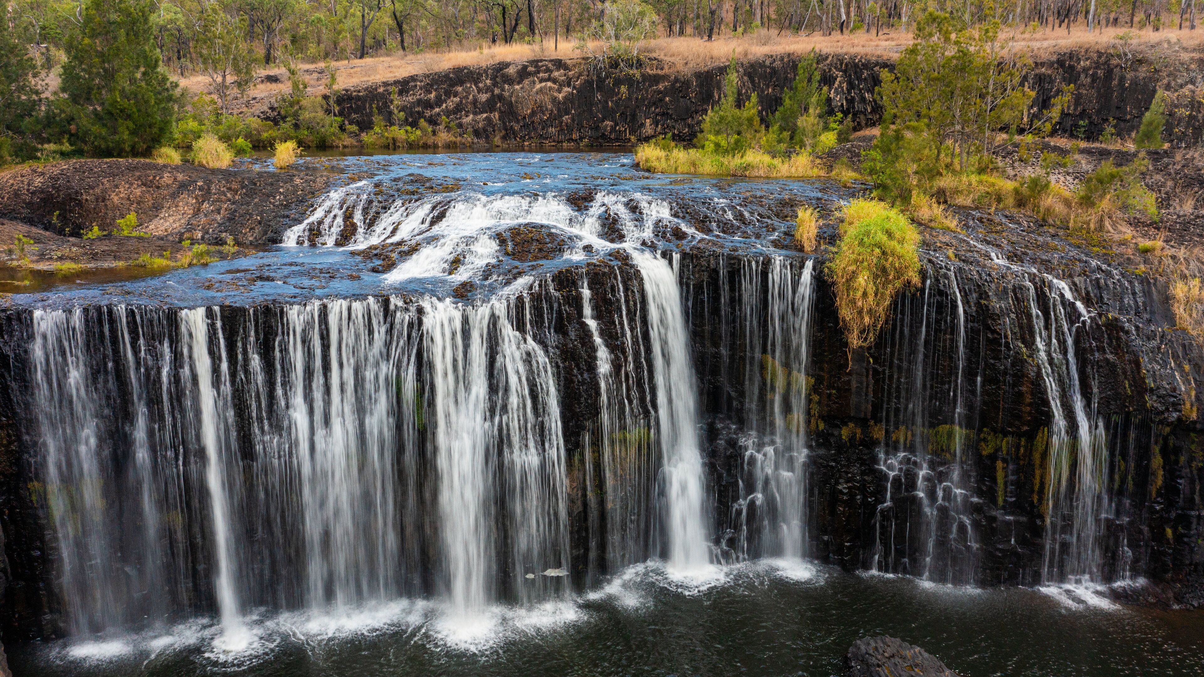 A waterfall cascading over a rocky cliff into a river, surrounded by sparse vegetation and trees.