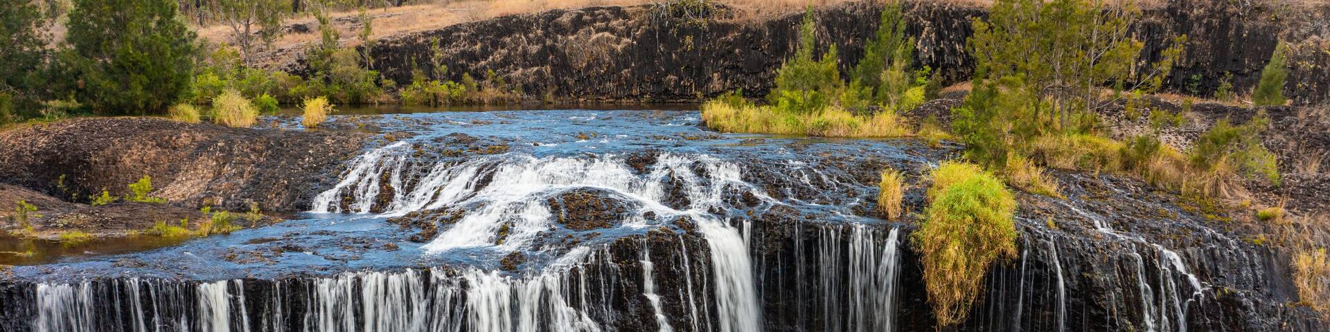 A waterfall cascading over a rocky cliff into a river, surrounded by sparse vegetation and trees.
