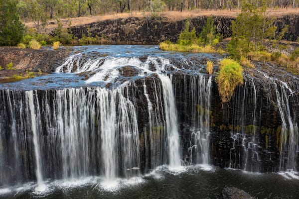 Ein Wasserfall stürzt über eine felsige Klippe in einen Fluss, umgeben von spärlicher Vegetation und Bäumen.