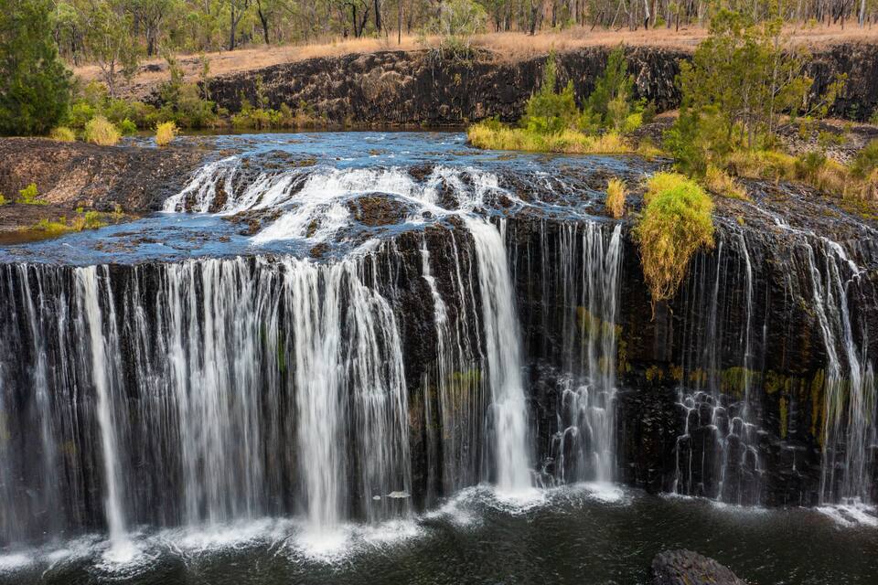 A waterfall cascading over a rocky cliff into a river, surrounded by sparse vegetation and trees.