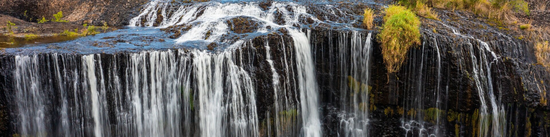 Uma cascata despencando sobre um penhasco rochoso em um rio, cercada por vegetação rala e árvores.