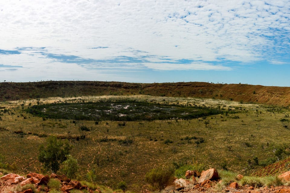 A vast, open landscape with a central circular formation surrounded by sparse vegetation and red rocky outcrops.