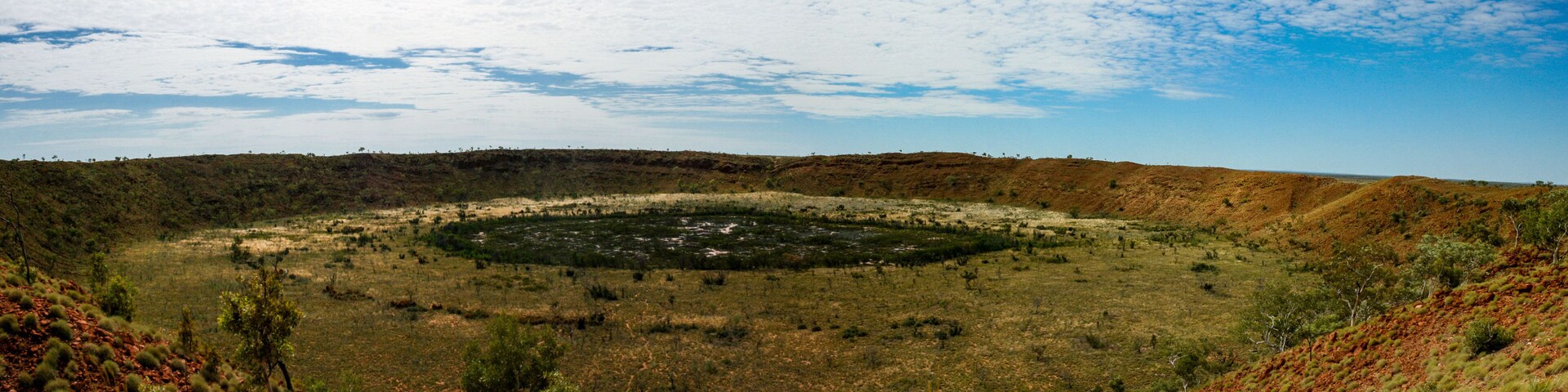 Uma extensa paisagem aberta com uma formação circular central, rodeada por vegetação rala e afloramentos rochosos vermelhos.