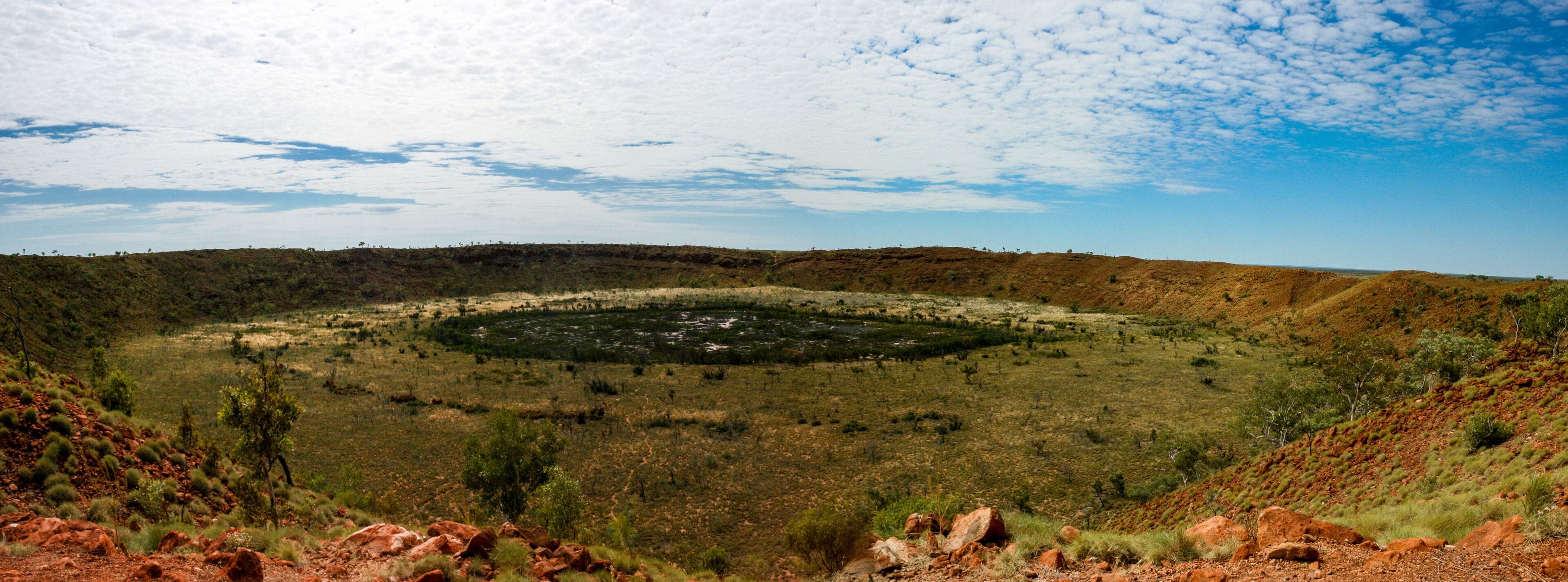 A vast, open landscape with a central circular formation surrounded by sparse vegetation and red rocky outcrops.