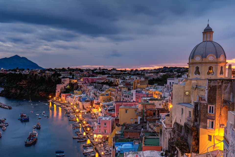 A coastal town at dusk with a prominent church, a mountain in the background, and numerous boats docked along the waterfront.