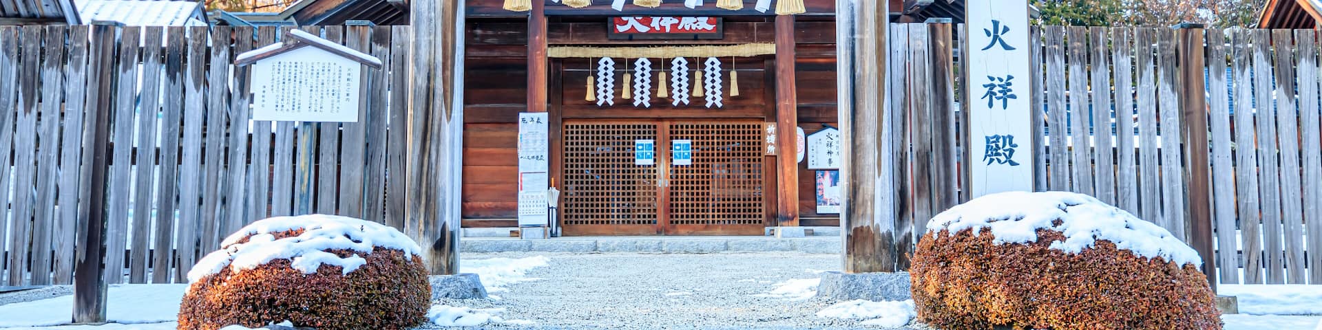 Un portón japonés de madera tradicional con arbustos cubiertos de nieve y una valla de madera al fondo.