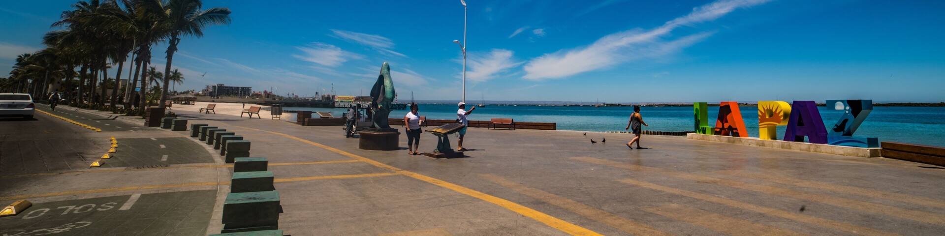 A coastal promenade with palm trees, benches, a sculpture, and colorful letters spelling 'LAPA' on the pavement.
