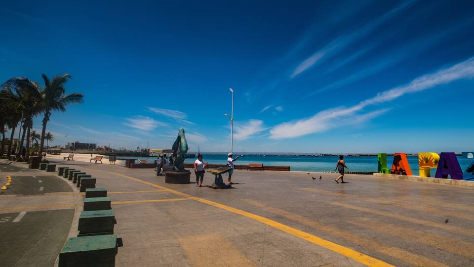 Un paseo marítimo con palmeras, bancos, una escultura y letras coloridas que deletrean 'LAPA' en el pavimento.