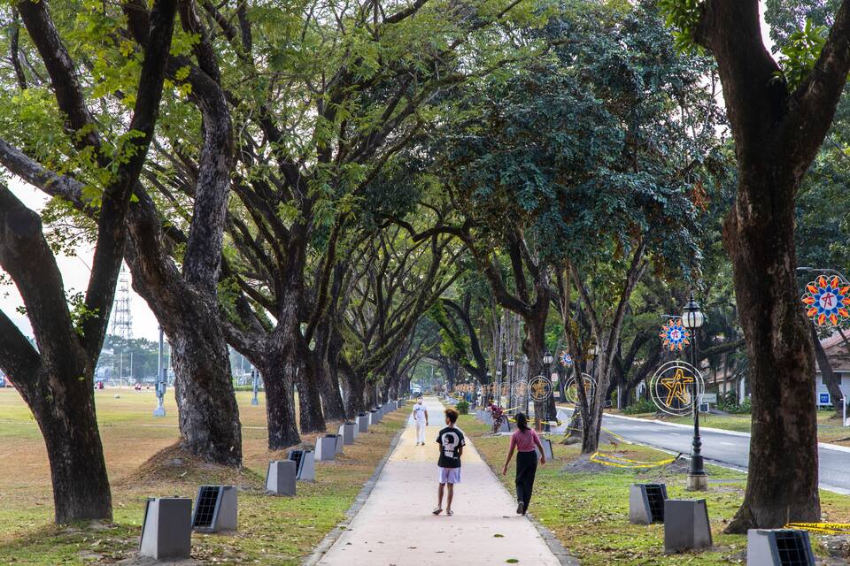 A tree-lined pathway with people walking, street lamps, and a road with a traffic sign.
