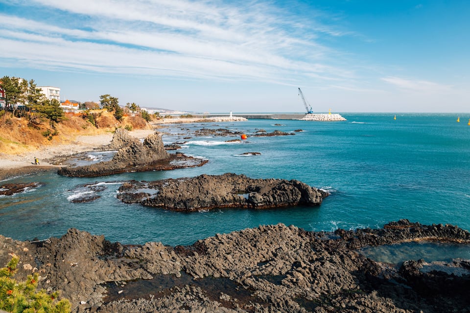 Une côte rocheuse bordée d’eaux d’un bleu limpide, un phare et une grue distante, visible sur une digue.
