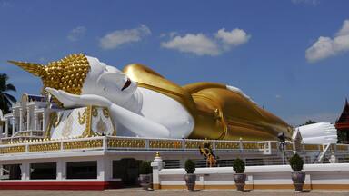 Une grande statue de Bouddha blanc et doré, en position allongée, ornée de détails complexes, se détachant sur un ciel bleu clair.