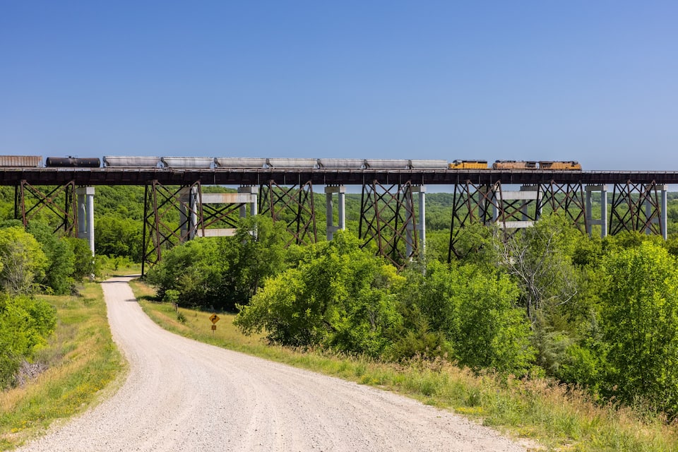 A gravel road leading to a railroad bridge with a train crossing it.