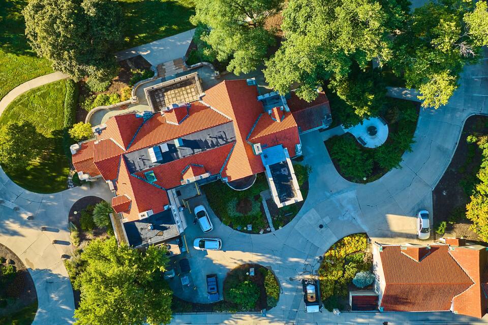 An aerial view of a residential area with a large house, multiple cars, and surrounding greenery.