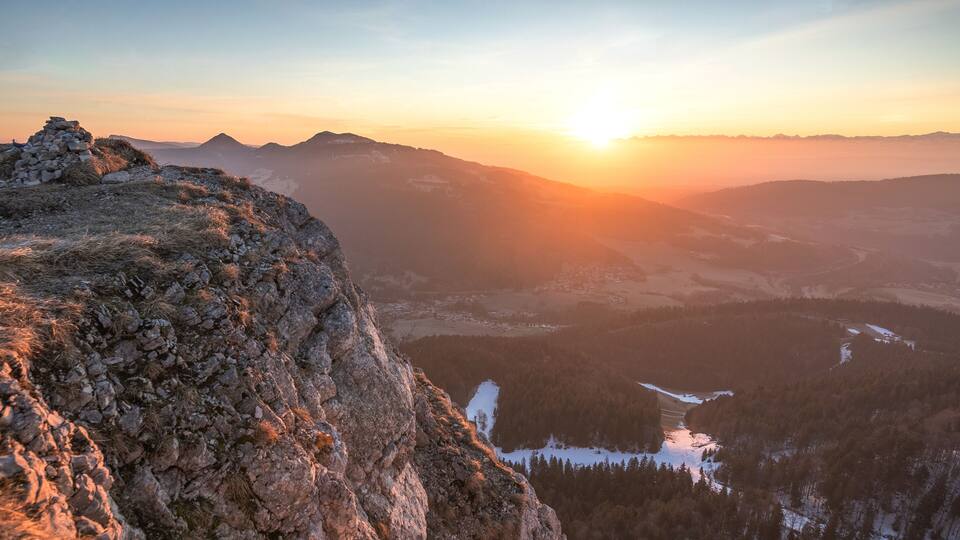 A mountainous landscape with a sunset, rocky terrain, and a valley with snow-covered areas.