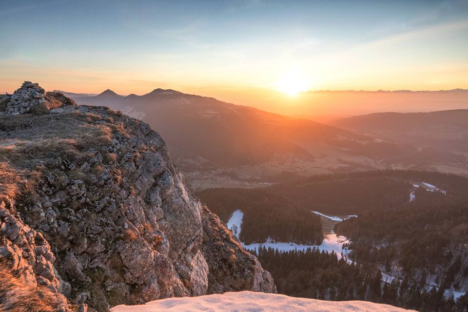 A mountainous landscape with a sunset, rocky terrain, and a valley with snow-covered areas.