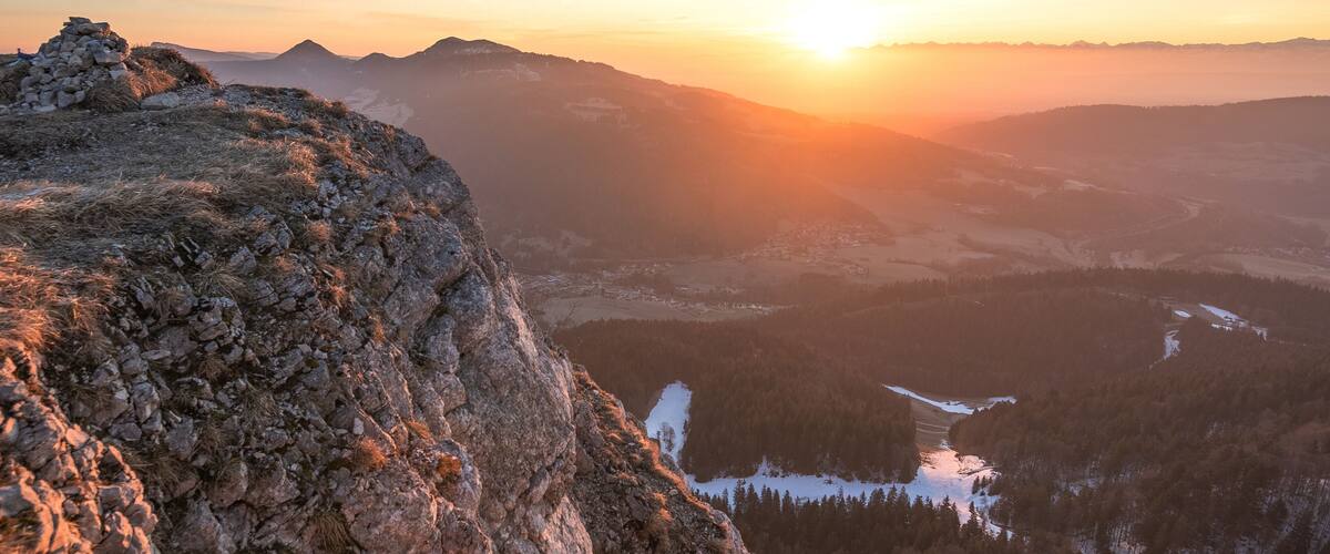 A mountainous landscape with a sunset, rocky terrain, and a valley with snow-covered areas.