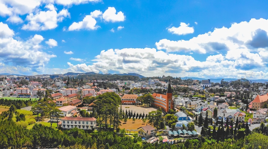 Paisagem urbana com uma torre de igreja imponente, uma grande praça aberta e um cenário montanhoso ao fundo.