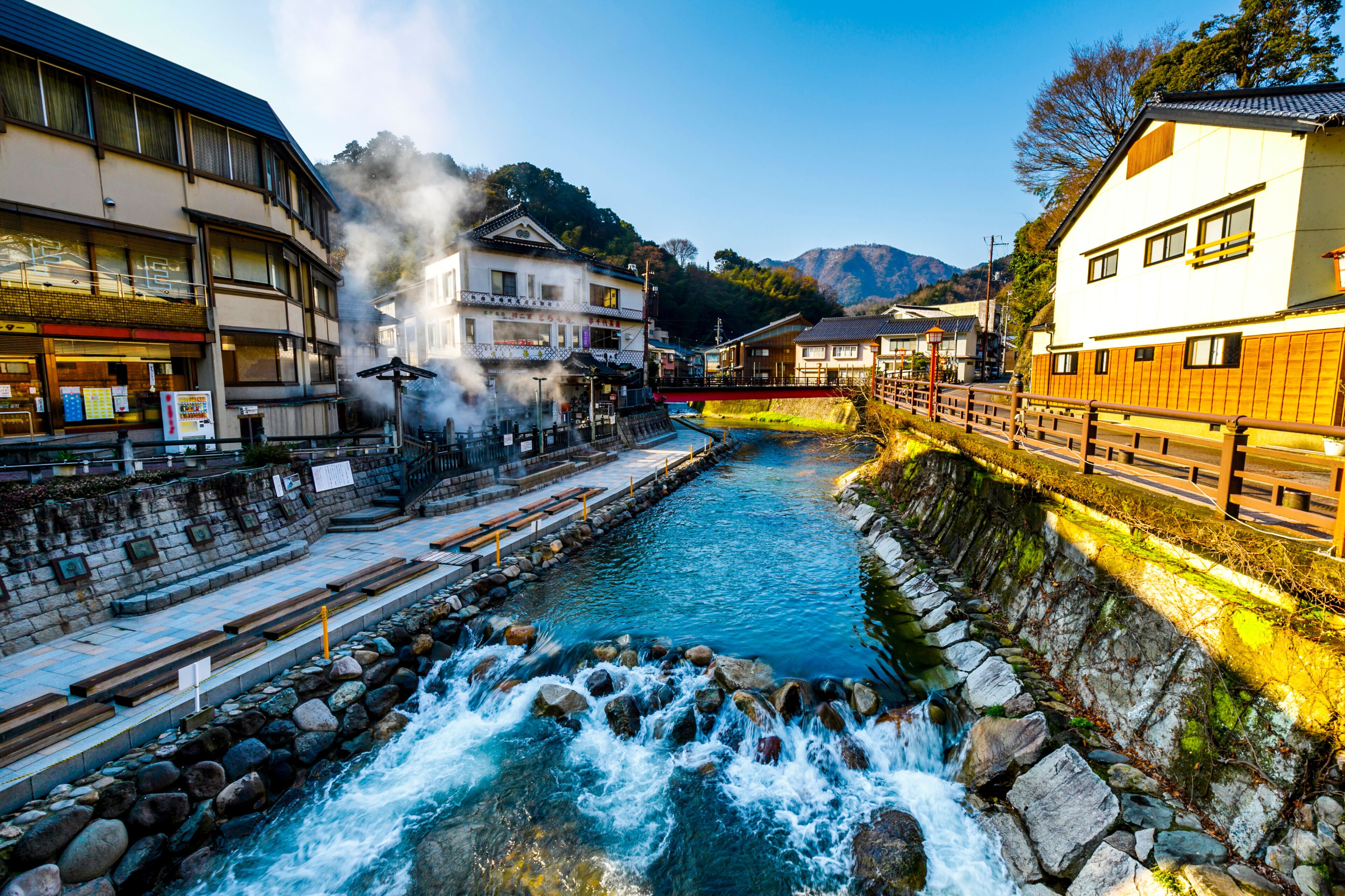 A river flowing through a town with traditional buildings, a bridge, and mountains in the background.