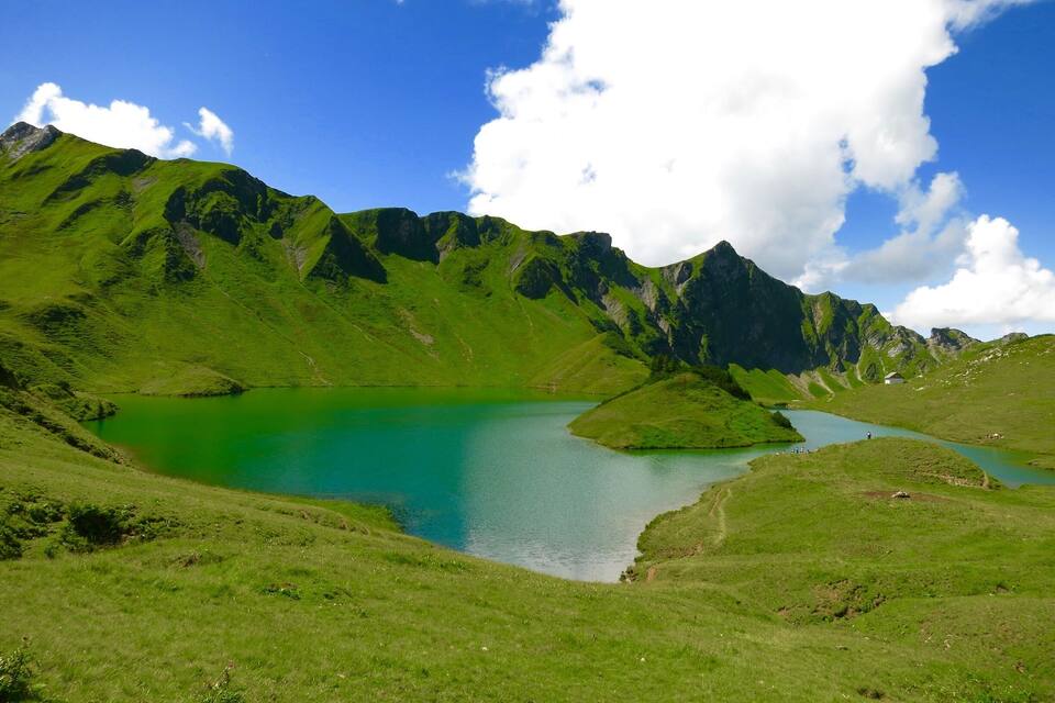 Un paisaje montañoso con un lago turquesa rodeado de colinas verdes y un pequeño edificio blanco.