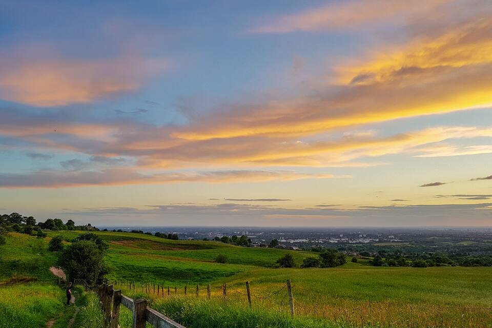 Un campo cubierto de hierba, con una cerca de madera y un horizonte urbano lejano bajo un cielo colorido.